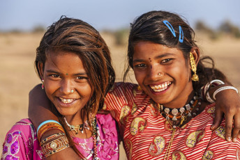 Two girls smiling and posing for the picture with their hands on each other’s shoulders wearing traditional dresses and ornaments in the Thar Desert of Jaisalmer, Rajasthan, India © Bartosz Hadyniak