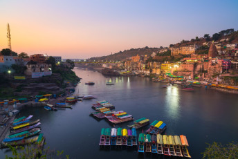 The sacred Omkareshwar Hindu Temple dominates the beautiful dusk cityscape scene with the Narmada River in the foreground. Dedicated to Lord Shiva, the name refers to the Lord of the Om Sound