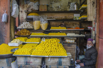 An old man is sitting in his local sweets and snacks shop containing ladoos and namkeen items at the local market in Madhya Pradesh, India. © Pete Burana