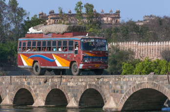 A passenger bus loaded with passengers and their luggage crosses a narrow bridge over the Betwa River while the magnificent Raj Palace is seen in the backdrop at Orchha, Madhya Pradesh, India. © Claudine Van Massenhove