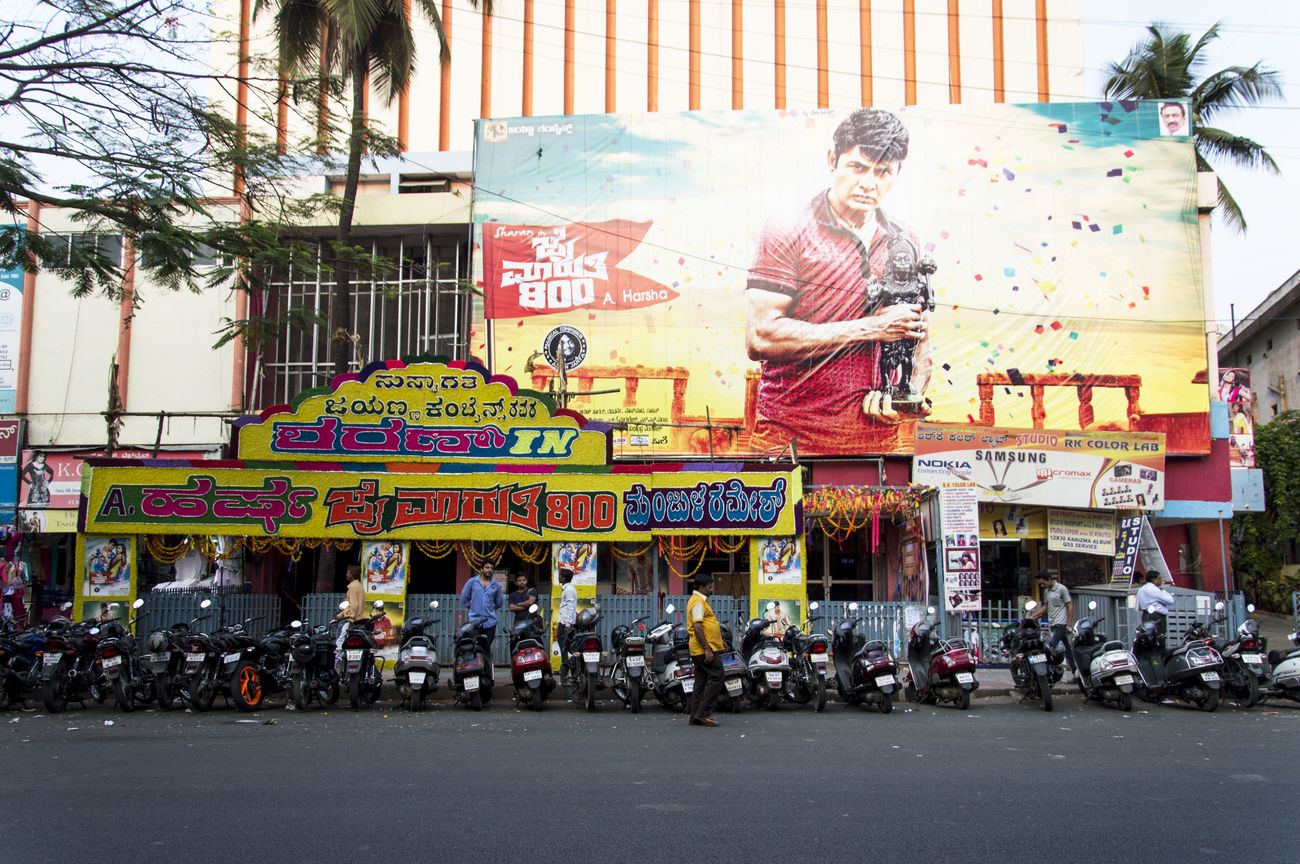 An enormous theater in Bangalore with a number of motorbikes parked in line queued to see a Kannada film