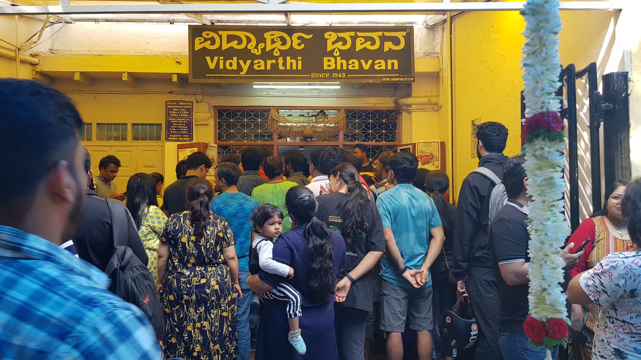 A large crowd of people waiting outside the well-known hotel 'Vidyarthi Bhavan' whose name is also written in local language 'Kannada'