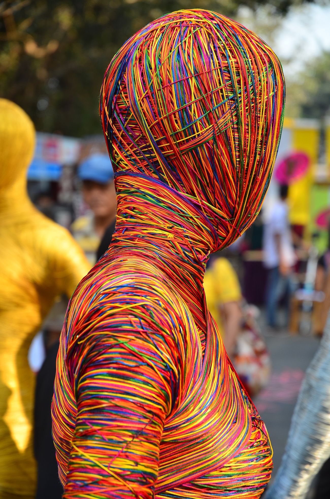 Human like objects wrapped up with multi colored threads stand on the outdoor ground at Kala Ghoda with a blurry image of unknown people at the backdrop 