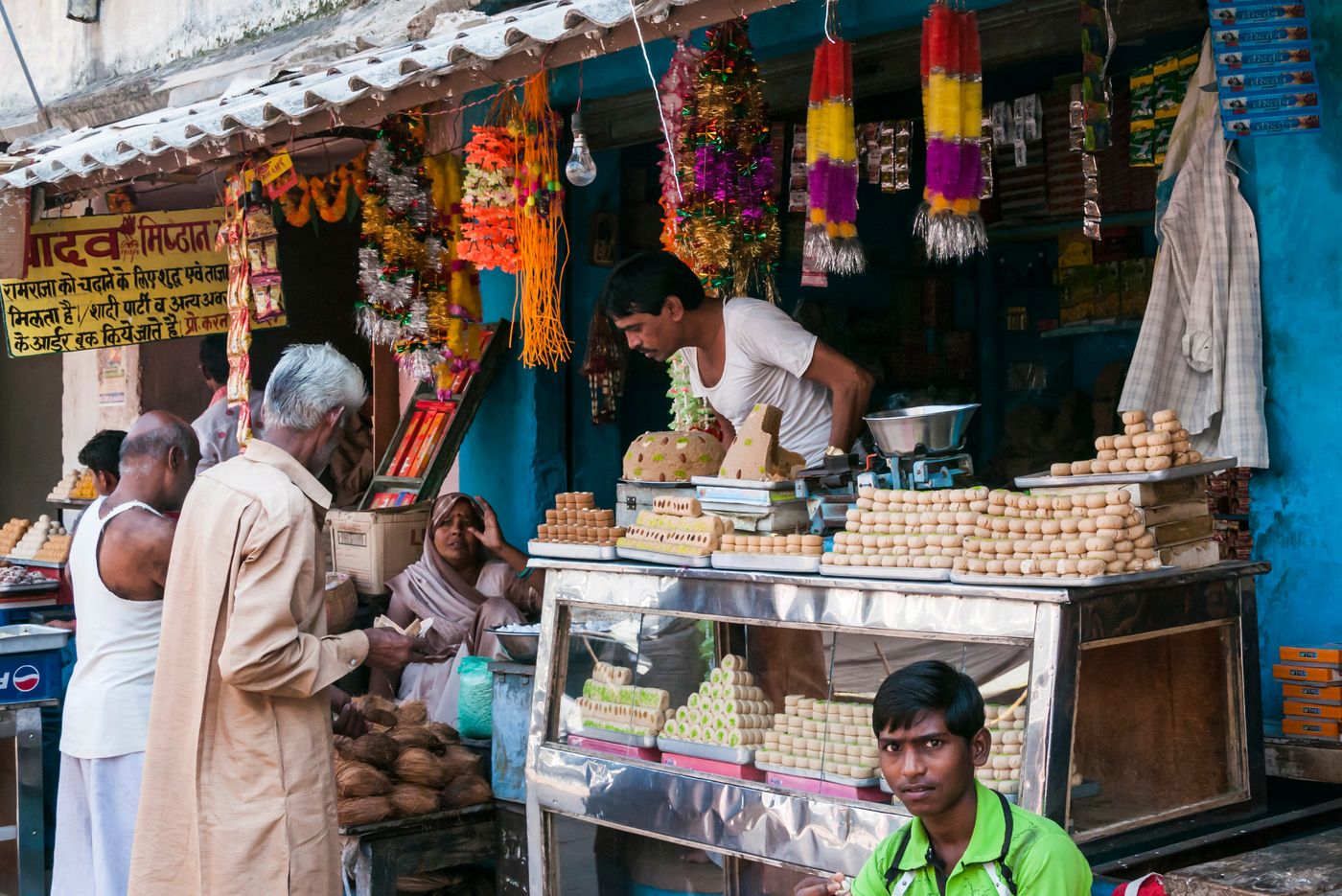 Wide variety of sweets for sale at this street stall. A woman sits in a chair observing the sale, Ram Raja Temple, Orchha