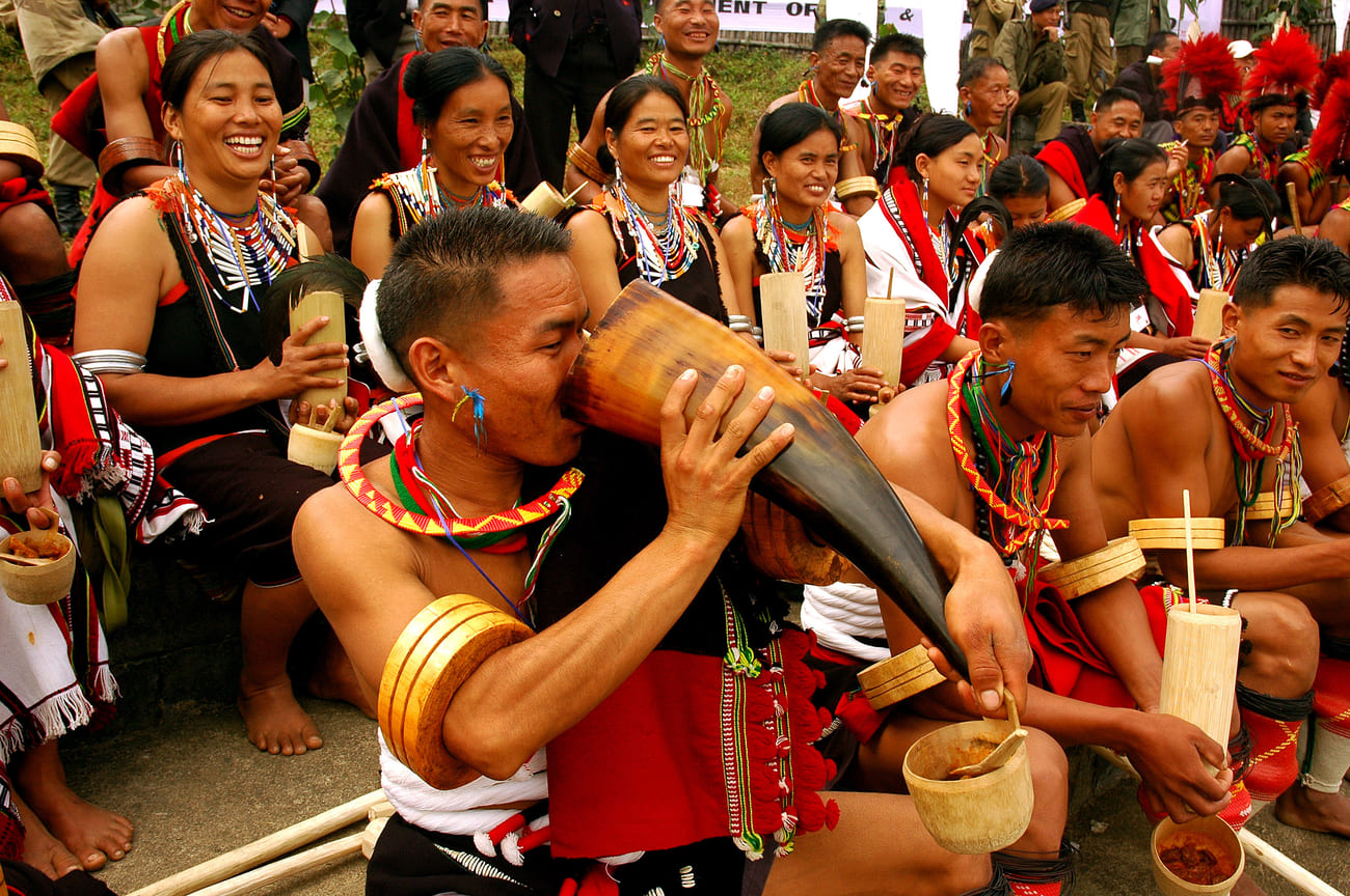 A man drinks from a huge horn, to the amusement of his fellow tribe members. Their traditional dress is an eye-catching profusion of colors