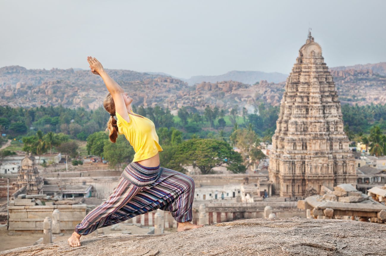 A woman practicing a yoga pose stretch on a rock looking over Hampi’s popular Virupaksha temple in Karnataka.