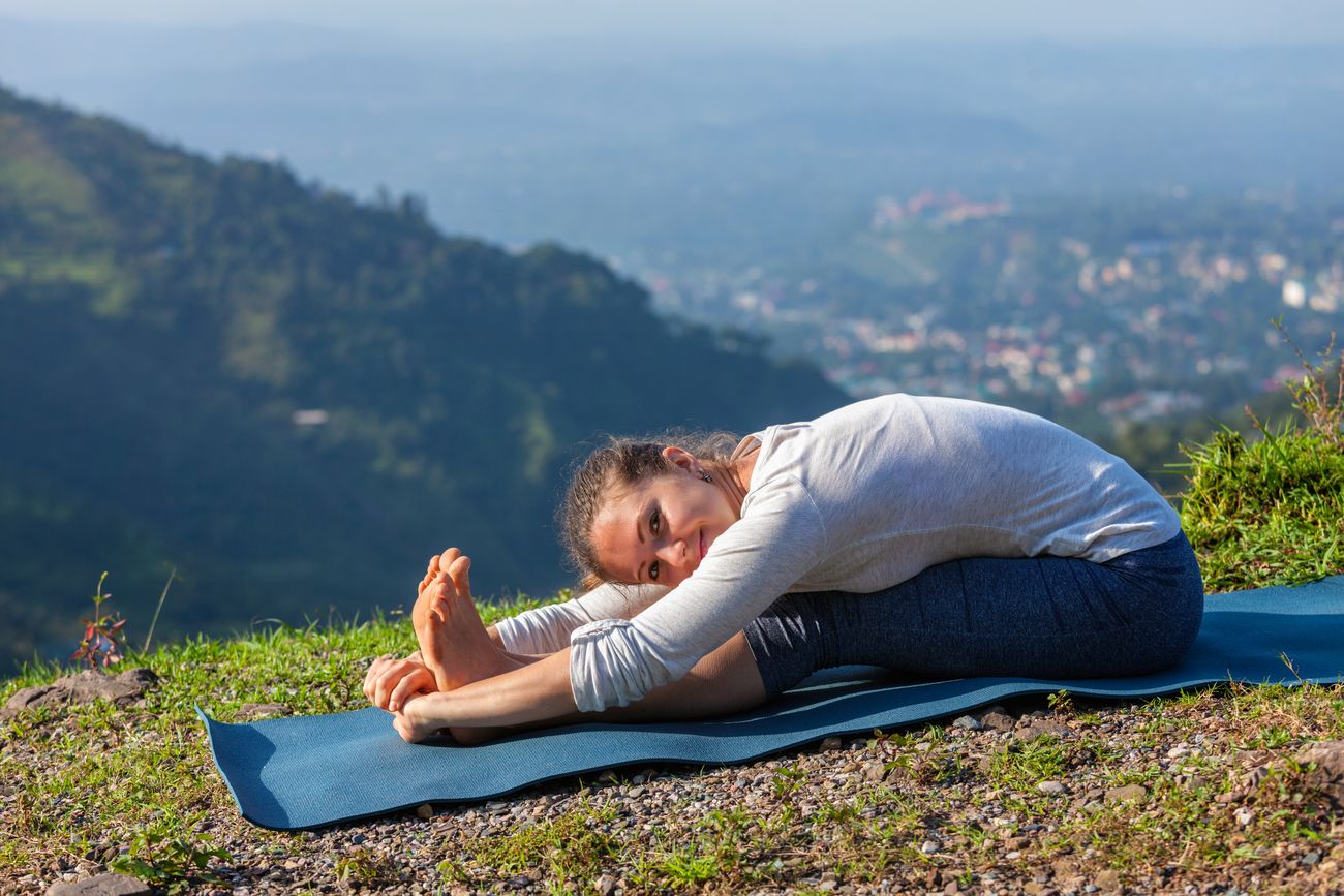 A young woman performs an asana from Ashtanga Vinyasa Yoga called Paschimottanasana seated amongst green mountains. Ashtanga Yoga requires synchronising one’s breathing with a continuous flow of postures. 