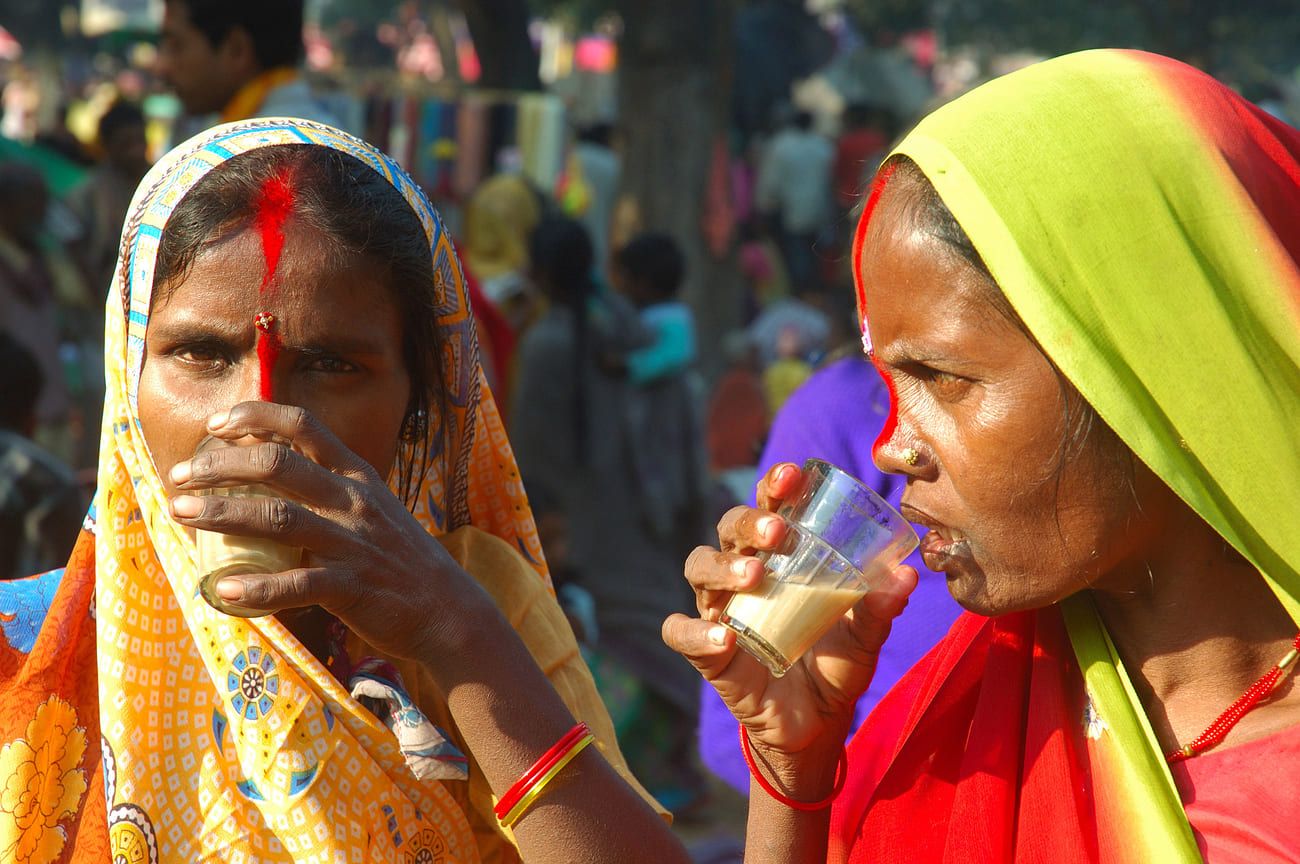 Close-up of two women, streaked with bright orange sindoor from nose tip to hairline, having sweet Indian milk tea