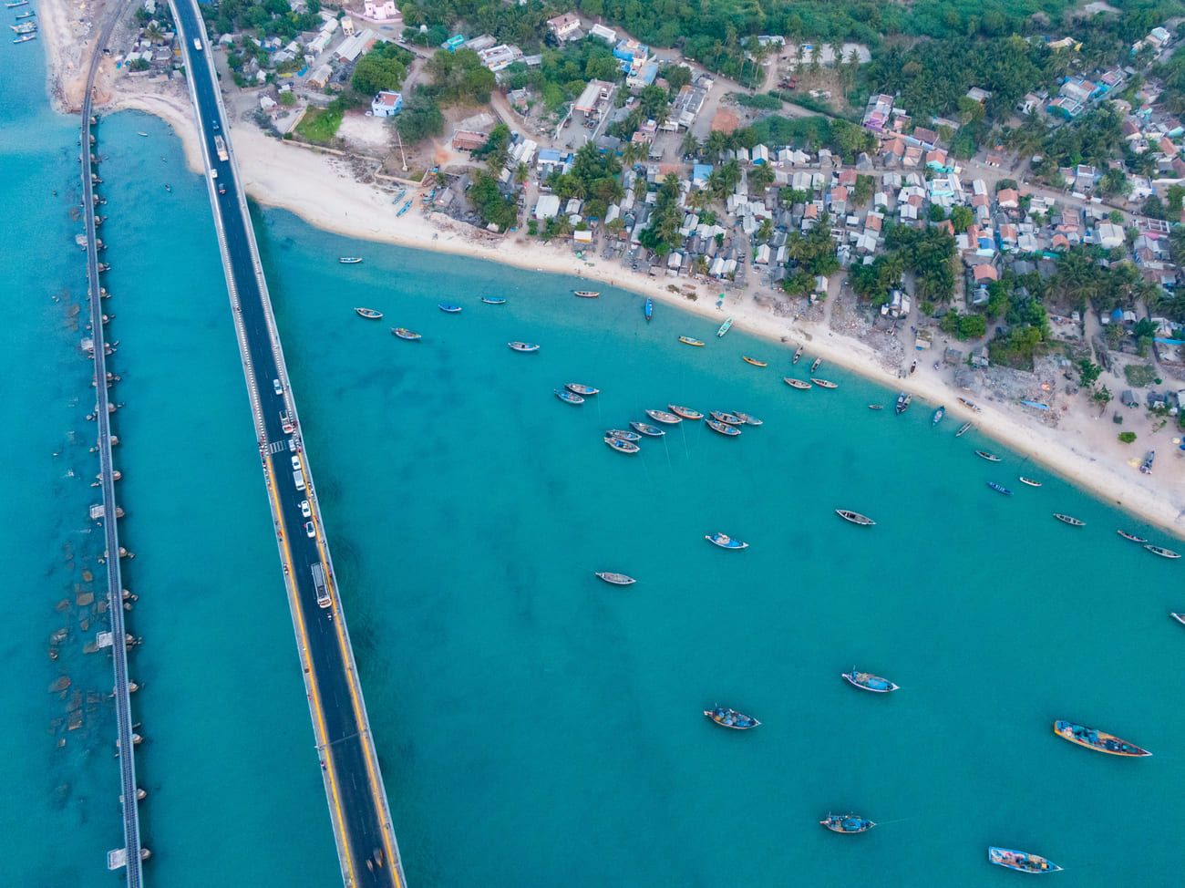 Spectacular drone shot of Pamban Bridge and road across a turquoise sea with boats floating near the beach 